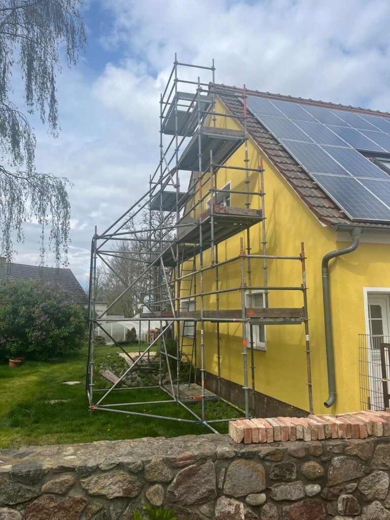 Das gelbe Haus mit Gerüst hat ein Solarpanel auf der Dachfläche, umgeben von grünem Garten.