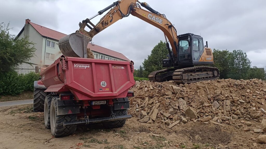 Ein gelber Bagger lädt Schutt in einen roten Anhänger auf einer Baustelle.