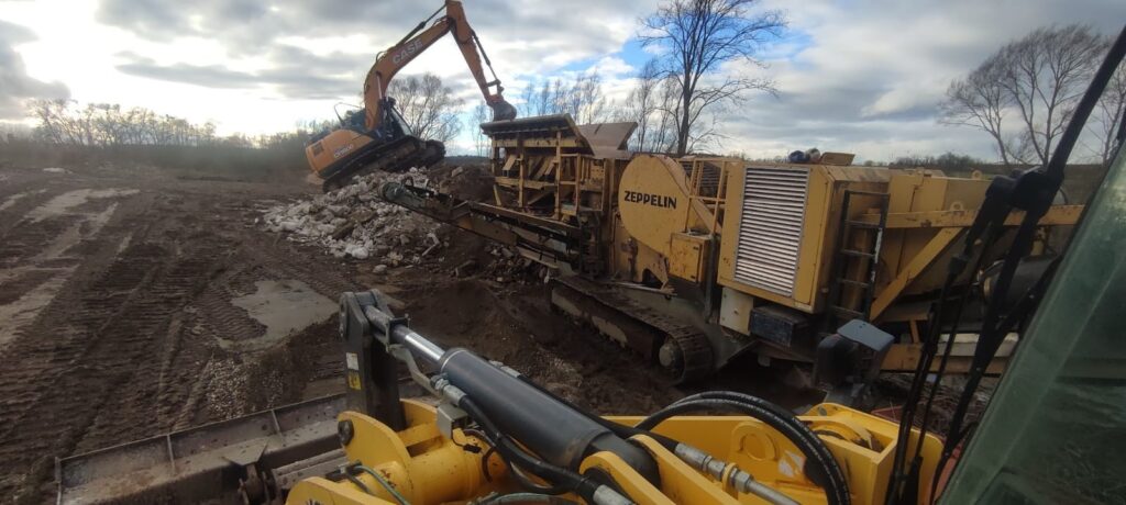 Eine Baustelle mit einem orangefarbenen Bagger, der Trümmer zu einer gelben Brechanlage führt, umgeben von Bäumen unter wolkigem Himmel