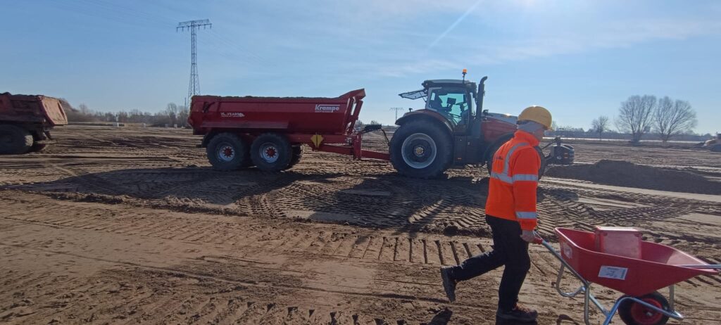 Eine Person mit orangener Jacke und Helm schiebt eine Schubkarre auf einer Baustelle entlang, während ein roter Traktor im Hintergrund steht.