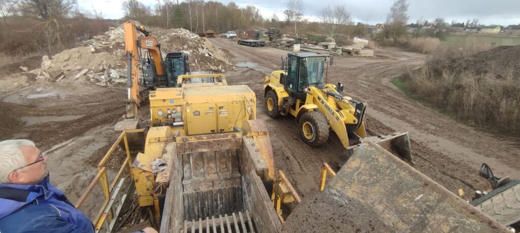 Ein Mann mit Brille und blauer Jacke beobachtet mehrere gelbe Baumaschinen auf einer schlammigen Baustelle.