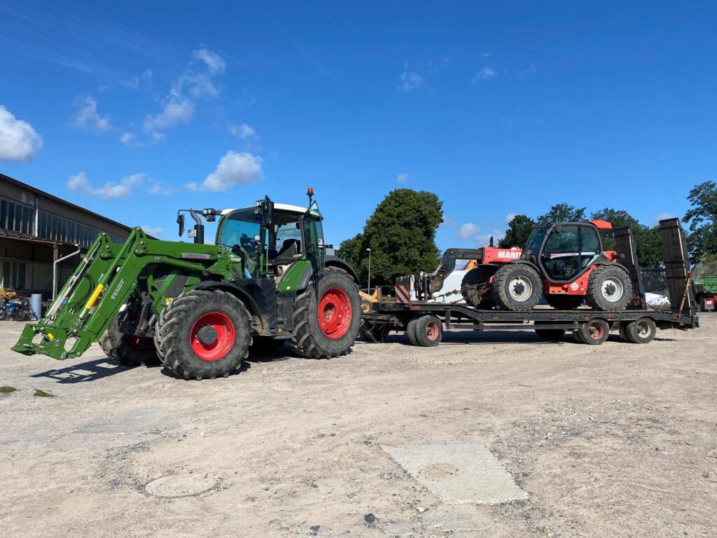 Ein grüner Traktor zieht einen Anhänger mit einem Kompaktlader auf einem sonnigen Bauernhof.